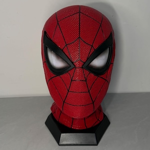 May include: A red Spider-Man mask featuring a textured surface with black web-patterned lines and large, white, mesh-covered eye lenses. The mask is mounted on a black, hexagonal display stand, showcasing a detailed, full-head design.