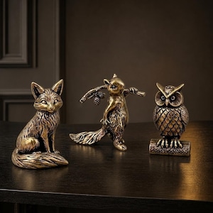 May include: Three metallic bronze-toned animal figurines featuring a seated fox with a bushy tail, a standing squirrel holding a branch with two pinecones, and an owl perched on a small log. Each piece displays detailed fur and feather textures.