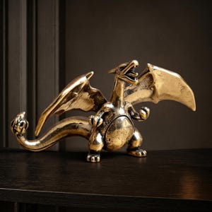 May include: A metallic gold-colored dragon figurine with outstretched wings, a long curved tail with a flame-like tip, and an open mouth. The creature stands in a bipedal stance with claws visible on its hands and feet, featuring a polished finish.
