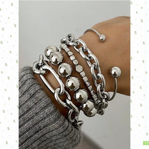 May include: A set of silver-toned bracelets featuring a variety of designs, including a thick oval link chain, a strand of large polished spheres, a square-link chain, and a thin open cuff bracelet with small spherical ends, worn on a wrist.