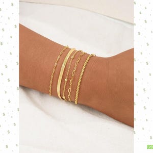 May include: A set of five gold-toned bracelets worn on a wrist. The collection includes a thin twisted chain, a smooth flat bangle, a delicate chain with small beads, a medium-sized oval link chain, and a thicker twisted rope chain bracelet.