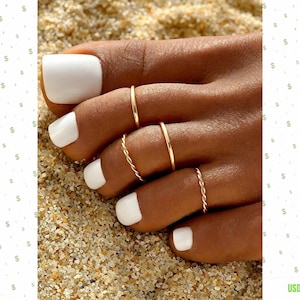 Puede incluir: Un conjunto de cuatro anillos para los dedos del pie en tono dorado, con dos bandas lisas y dos bandas de estilo cuerda trenzada. Las uñas están pintadas con esmalte blanco sólido y las joyas se muestran sobre un fondo de arena texturizada.