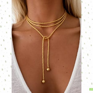 Könnte beinhalten: Eine goldfarbene Lariat-Halskette mit einem mehrsträngigen Ketten-Design und einer strukturierten, gewebten Optik. Die Kette wird um den Hals gewickelt und mit einem zentralen Knoten fixiert, wobei zwei herabhängende Enden in kleinen, glatten Kugeln enden.