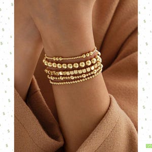 Puede incluir: Una pila de cinco pulseras de cuentas doradas en la muñeca. Las pulseras presentan cuentas de diversos tamaños y formas, incluyendo esferas redondas lisas y pequeñas cuentas planas en forma de disco, creando un aspecto metálico superpuesto.