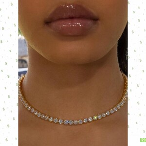 Può includere: Una collana tennis color oro caratterizzata da una fila continua di pietre rotonde, trasparenti e sfaccettate incastonate in una catena metallica. La collana poggia sul collo, mostrando un design uniforme di gemme circolari scintillanti.