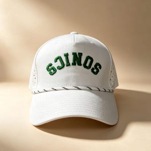 K&ouml;nnte beinhalten: Eine wei&szlig;e Baseballkappe mit dem Wort "SONICS", das in dunkelgr&uuml;nen Blockbuchstaben auf der Vorderseite eingestickt ist. Die Kappe verf&uuml;gt &uuml;ber eine dekorative wei&szlig;e Kordel mit feinen schwarzen Akzenten an der Basis der Krone sowie perforierte Seitenteile.