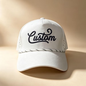 May include: A white baseball cap featuring the word Custom in dark navy blue cursive embroidery on the front panel. The cap includes a white and navy blue twisted rope detail across the base of the crown and perforated side panels for ventilation.
