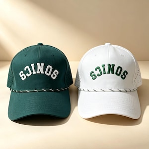 Op de afbeelding: Twee baseballpetten, één donkergroen en één wit, elk met het woord SONICS in een contrasterende kleur op de voorkant geborduurd. Beide petten zijn voorzien van een decoratief gevlochten koord aan de basis en geperforeerde zijpanelen.