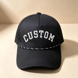 May include: A black baseball cap featuring the word CUSTOM embroidered in white capital letters across the front panels. The cap includes a black and white braided rope detail across the base of the crown and a curved brim with stitching.