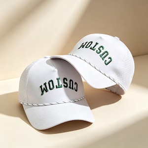 Könnte beinhalten: Zwei weiße Baseballkappen mit dem Wort CUSTOM, das in dunkelgrünen Großbuchstaben auf die Vorderseite gestickt ist. Jede Kappe verfügt über eine dekorative, gedrehte schwarz-weiße Kordel am unteren Rand der Krone und perforierte Seitenteile.