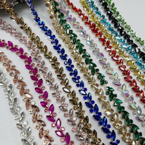 Puede incluir: Múltiples tiras de adorno decorativo con pedrería de corte marquesa dispuesta en un patrón repetitivo de hojas. El adorno está disponible en varios colores, incluyendo transparente, rosa, azul, verde, rojo, dorado y negro, con engastes metálicos.