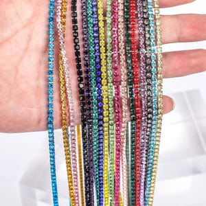 May include: A collection of multiple long, thin strands of rhinestone chain in a variety of vibrant colours including blue, yellow, pink, black, purple, green, and red. Each strand features small, uniform, faceted crystals set in metal.