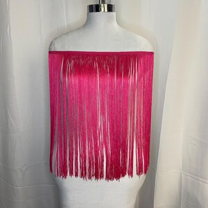 May include: A vibrant hot pink off-the-shoulder top featuring a straight neckline and long, flowing fringe that extends from the chest down to the waist. The garment is displayed on a mannequin, showcasing the uniform vertical strands of fringe.