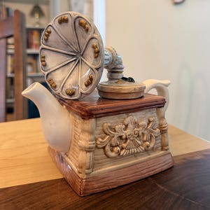 May include: A ceramic teapot shaped like a vintage gramophone with a cream-colored body, a brown wood-grain base, and a flared horn top. The teapot features a white spout, a handle, and intricate relief carvings on the side panels with gold accents.