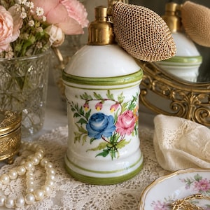 May include: A white ceramic perfume bottle with a gold-toned metal atomizer pump and a gold mesh bulb. The bottle features hand-painted floral motifs including blue and pink roses with green leaves and thin green bands around the top and base.