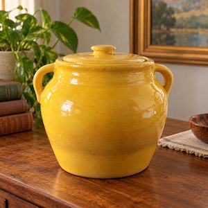 May include: A bright yellow ceramic jar with a glossy finish, featuring two side handles and a matching lid. The jar has a rounded, bulbous body and a wide opening, showcasing a smooth, solid-colored surface with subtle variations in the yellow glaze.