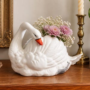 May include: A white ceramic swan-shaped planter featuring a glossy finish, a bright orange beak, and detailed molded feather textures. The swan is filled with a small bouquet of pink chrysanthemums and white baby's breath flowers.