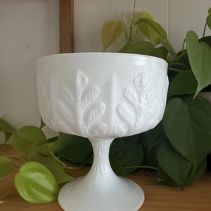 Peut inclure: Une coupe à pied en verre opalin blanc présentant un motif en relief de feuilles de fougère sur l'extérieur du bol et un design texturé rappelant l'écorce d'arbre sur la base. Le verre possède une finition opaque et lisse sur toute la pièce.