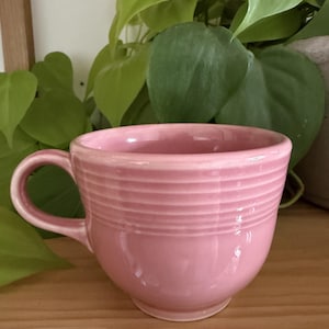 Könnte beinhalten: Ein einfarbiger, hellrosa Keramikbecher mit einer Reihe horizontaler, geprägter Ringe im oberen Bereich des Körpers. Der Becher verfügt über einen geschwungenen Henkel auf der linken Seite und eine glatte, glänzende Oberfläche.