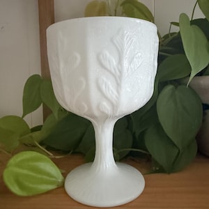 Peut inclure: Un calice en verre opalin blanc présentant un motif de feuilles en relief sur le bol. La tige est cannelée et repose sur une base circulaire lisse. Le verre possède une finition opaque et laiteuse avec une texture issue du moulage.