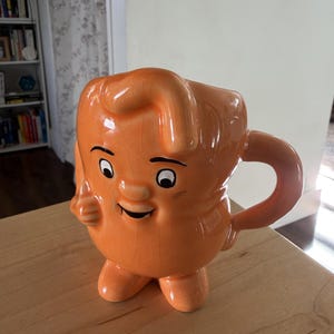 Op de afbeelding: Een glanzende oranje keramische mok in de vorm van een stripfiguur met een lachend gezicht, zwarte ogen en wenkbrauwen. De mok heeft een textuur met craquelé glazuur, een gebogen handvat aan de rechterkant en twee kleine voetjes aan de basis.