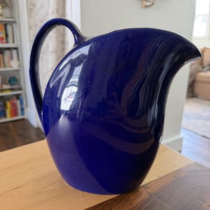 May include: A deep cobalt blue ceramic pitcher with a glossy finish, featuring a curved handle on the left side and a wide, prominent pouring spout on the right. The vessel has a smooth, rounded body and a wide base, reflecting light on its surface.