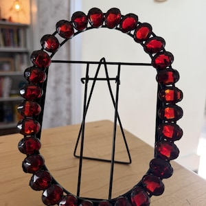 May include: An oval-shaped black metal picture frame featuring a border of faceted, deep red circular glass stones. The frame is supported by a thin, black wire easel stand attached to the back, allowing it to stand upright on a flat surface.