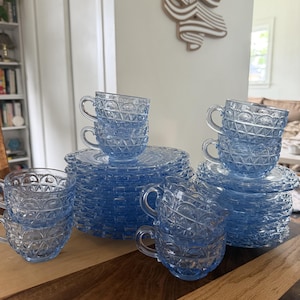 Op de afbeelding: Een set lichtblauwe glazen theekopjes en bijpassende schoteltjes met een gestructureerd geometrisch patroon van herhalende bogen en ruitvormen. Het doorschijnende blauwe glaswerk is in meerdere stapels gerangschikt.