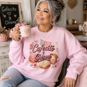 May include: A light pink long-sleeve sweatshirt featuring a graphic print with the text Cafecito y Chisme, two concha sweet breads, a floral coffee mug, and decorative bunting, surrounded by small pink flowers and hearts.