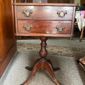 May include: A dark brown wooden pedestal table featuring two stacked drawers with ornate brass-toned metal pulls. The table is supported by a central turned column that branches into three curved legs with claw-style feet at the base.
