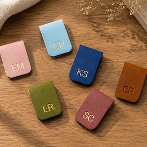 May include: Six rectangular magnetic bookmarks in various colors including light blue, pink, dark blue, olive green, brown, and maroon. Each bookmark features two gold-embossed capital letters: EM, CP, KS, CR, LR, and SC, centered on the front surface.