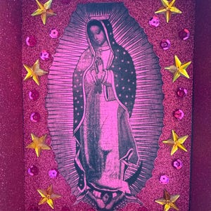 May include: A pink glitter box featuring an image of the Virgin Mary. The image is surrounded by a pink oval border and adorned with gold star-shaped embellishments and pink sequins. The box has a glittery finish.