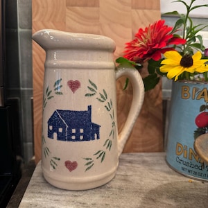Peut inclure: Une cruche en céramique beige ornée d'une illustration de maison bleue au centre d'une couronne de feuilles vertes, agrémentée de deux petits cœurs rouges au-dessus et en dessous. La cruche possède une anse robuste et une finition mouchetée.