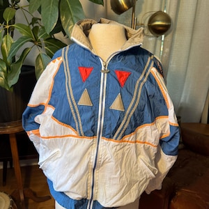 May include: A blue, white, and tan windbreaker jacket featuring a full-length front zipper, a high collar, and two side pockets. The chest is decorated with two red triangles and two tan triangles, accented by thin orange piping and tan trim details.