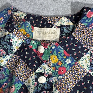 May include: A Sportalm Kitzbühel blouse featuring a patchwork design of various floral patterns in navy blue, red, yellow, green, and white. The garment has a round neckline, a front button placket with white embossed buttons, and a fabric label.