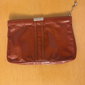 May include: A rectangular rust-colored leather clutch bag featuring a smooth finish, a vertical central panel with two parallel stitched lines, a silver-toned metal rectangular accent at the top center, and a silver-toned chain pull on the zipper.