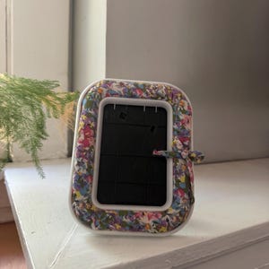 以下が含まれることがあります： マルチカラーの花柄の布で覆われた白いプラスチック製の縁を持つ長方形のフォトフレーム。フレームには黒いプラスチック製のインサートが含まれており、右側には装飾的なディテールとして小さな布製の蝶結びが結ばれています。