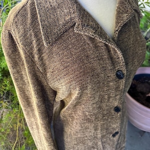 May include: A long-sleeved button-down jacket in a textured brown fabric with a subtle black speckled pattern. The garment features a classic collar, long sleeves, and three dark buttons down the front, displayed on a mannequin.