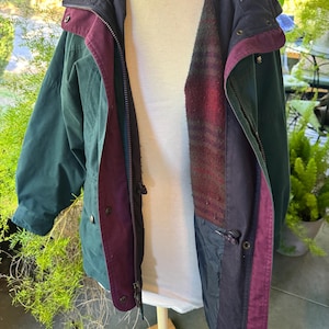 May include: A multi-layered jacket featuring a dark green outer shell, a deep purple inner placket, and a dark navy blue collar. The interior lining displays a textured, dark red and black plaid pattern. The jacket includes a full-length zipper.
