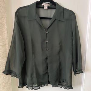 May include: A sheer, dark olive green button-down shirt with a pointed collar, three-quarter length sleeves, and ruffled cuffs. The garment features a ruffled hemline and a front button closure. A label reading "notations" is visible at the neck.