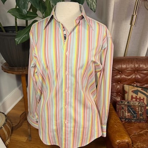 May include: A long-sleeve button-down shirt featuring a vertical striped pattern in shades of yellow, pink, light blue, and white. The shirt has a pointed collar, a front button placket, and a dark patterned fabric lining visible inside the collar.
