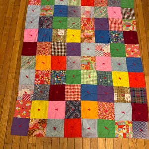 May include: A rectangular patchwork quilt composed of numerous multi-colored fabric squares in various patterns including gingham, paisley, floral, and solid colors. Each square is tied in the center with a small red yarn knot for texture.