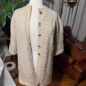 May include: A beige, textured knit cardigan featuring a round neckline, elbow-length sleeves, and a front button closure with five round, light-colored buttons. The garment is displayed on a mannequin, showcasing a fuzzy, looped yarn construction.