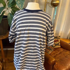 Puede incluir: Una camiseta de manga corta con un patrón de finas rayas horizontales en azul marino y blanco. La prenda tiene un cuello redondo en azul marino sólido y un corte relajado, exhibida sobre un torso de maniquí para mostrar el diseño.