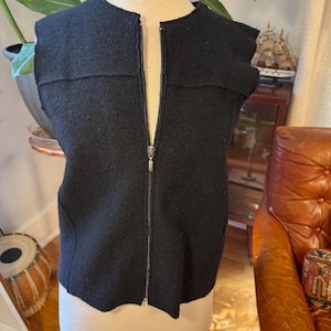 Puede incluir: Un chaleco negro sin mangas con cierre de cremallera frontal completo y un tirador de metal plateado. La prenda presenta un cuello redondo y detalles de costuras horizontales en la parte superior del pecho, confeccionada en un tejido texturizado.
