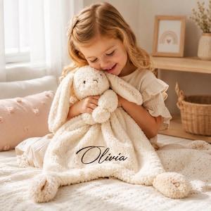 Puede incluir: Una manta de seguridad de felpa color crema con una cabeza de conejo de peluche con orejas largas y caídas y un cuerpo de textura suave. La tela está bordada con el nombre Olivia en una letra cursiva de color marrón oscuro en el frente.
