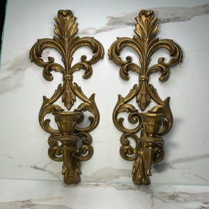 Könnte beinhalten: Ein Paar passende goldfarbene Wandleuchter mit einem verzierten Fleur-de-Lis-Design. Jeder Leuchter verfügt über einen zentralen Kerzenhalter, der von einem dekorativen Schnörkelrahmen mit filigranen blattartigen Details und einer metallischen Oberfläche getragen wird.