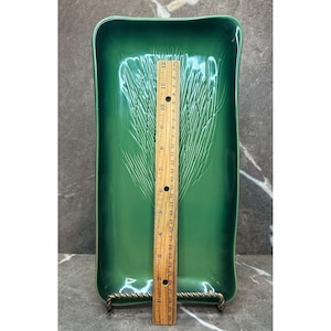 Könnte beinhalten: Ein rechteckiges grünes Keramiktablett mit einem geprägten Weizenährenmuster. Ein Holzlineal liegt vertikal in der Mitte und zeigt eine Länge von etwa 30 cm. Das Tablett hat eine glänzende Oberfläche und leicht erhöhte Ränder.