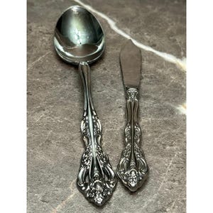 May include: A silver-toned metal spoon and a matching butter knife featuring intricate, embossed floral and scrollwork patterns on the handles. Both pieces have a polished, reflective finish and a detailed, ornate design on the stems.