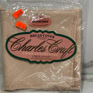 May include: A square piece of beige Charles Craft cross stitch fabric measuring 18 inches by 18 inches, packaged in a clear plastic bag. The label reads Bread Cover, Soft Touch, 14 Count, and E-Z Care Dacron/Polyester.